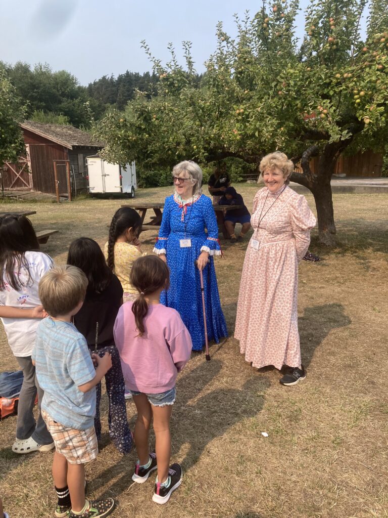 A tour group for Funhouse youth is led by volunteers Fran and Cheryl. They are gathered in the apple orchard outside the Museum.